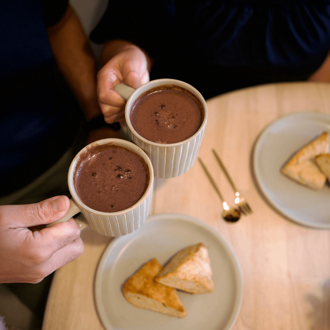 
                  
                    Holding two cups of Sana Vidal hot cocoa over table with savory scones
                  
                